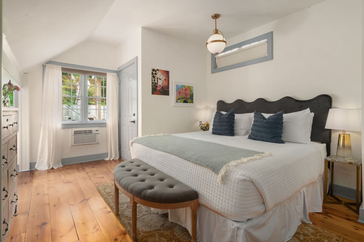 A bedroom with a large bed, a bench, a window with curtains, and a wall-mounted lamp.