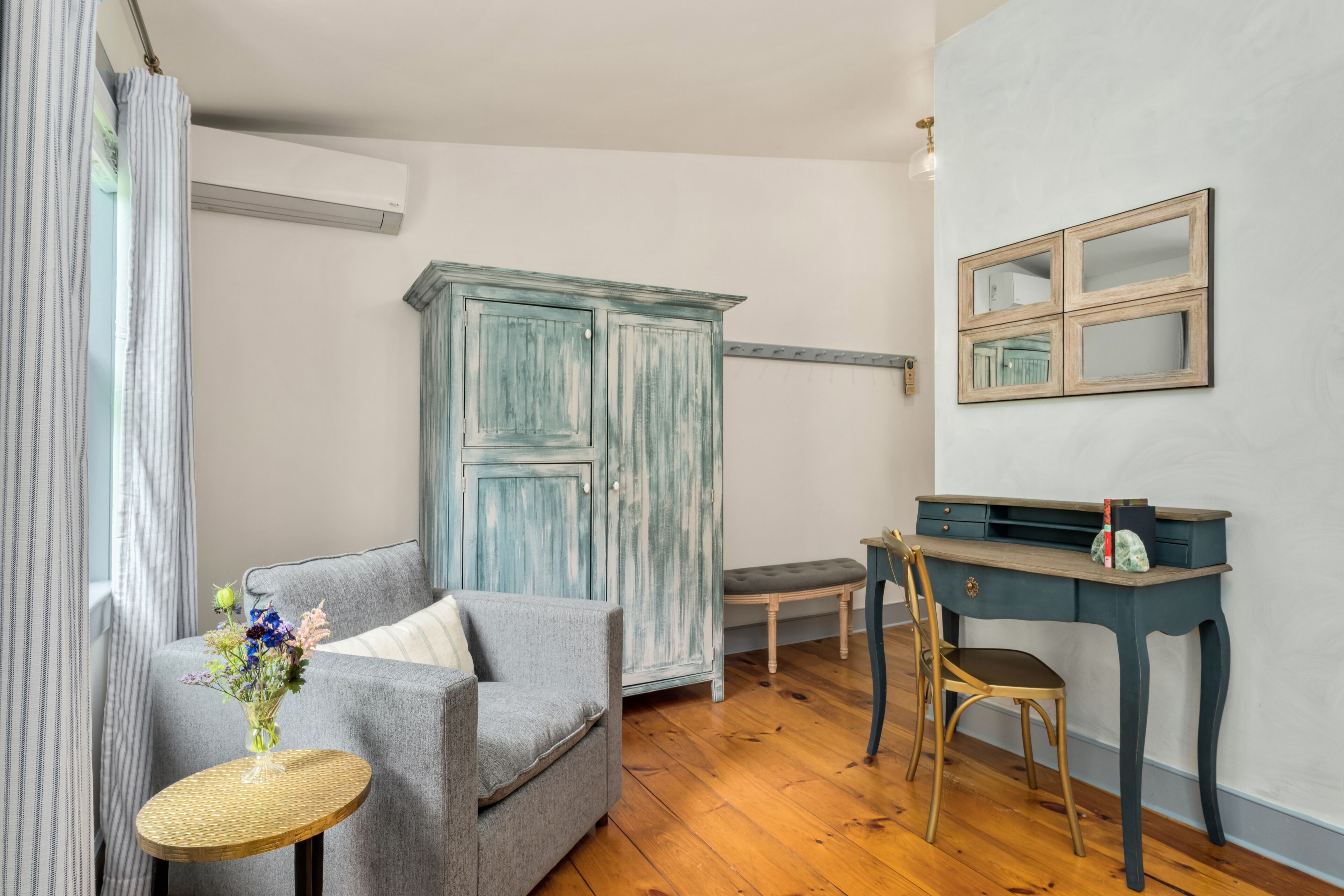 Una habitación con un sofá gris, una mesita redonda con un florero, una silla de madera, un escritorio con un espejo y un armario azul.