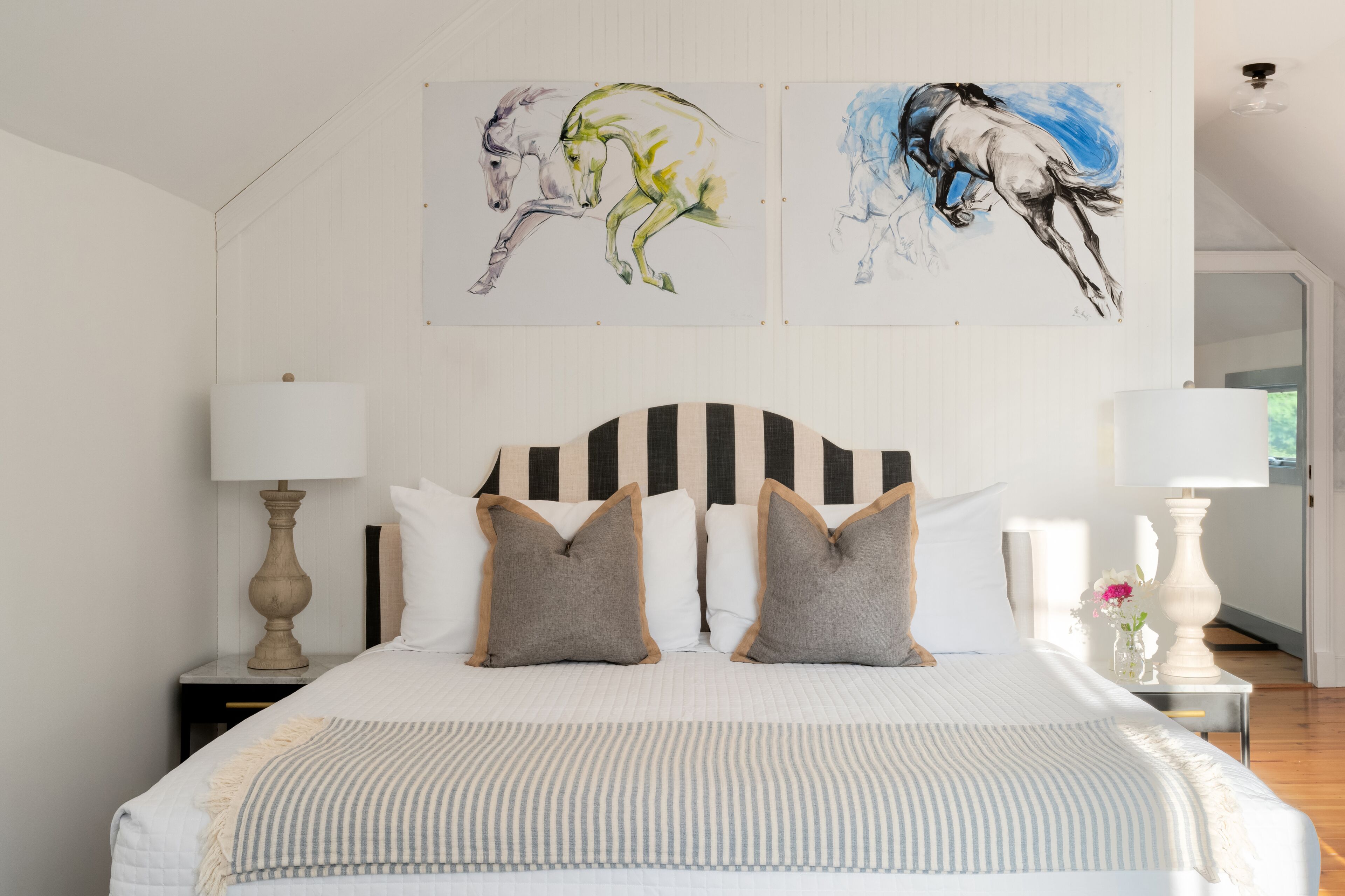 A bedroom with a bed, two bedside lamps, a headboard, and two paintings on the wall.