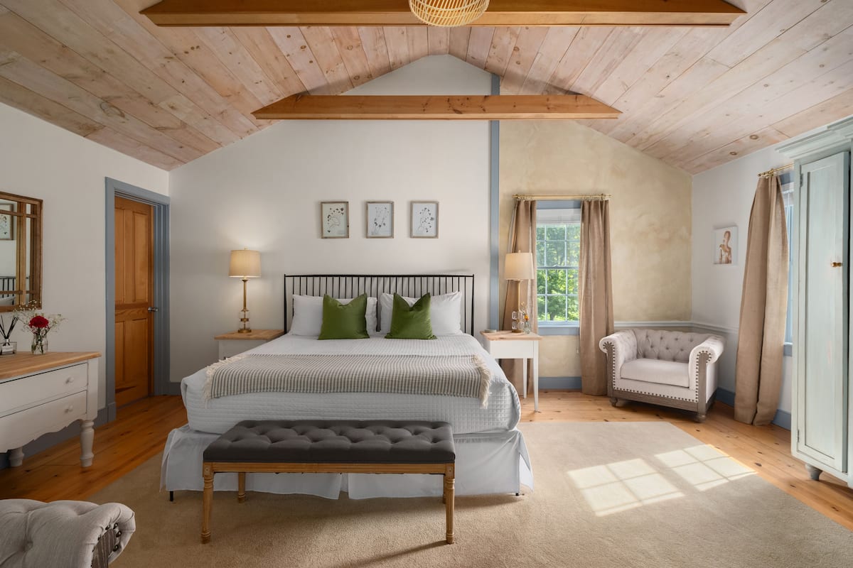 A bedroom with a large bed, a bench, a chair, a nightstand, and a window with curtains.
