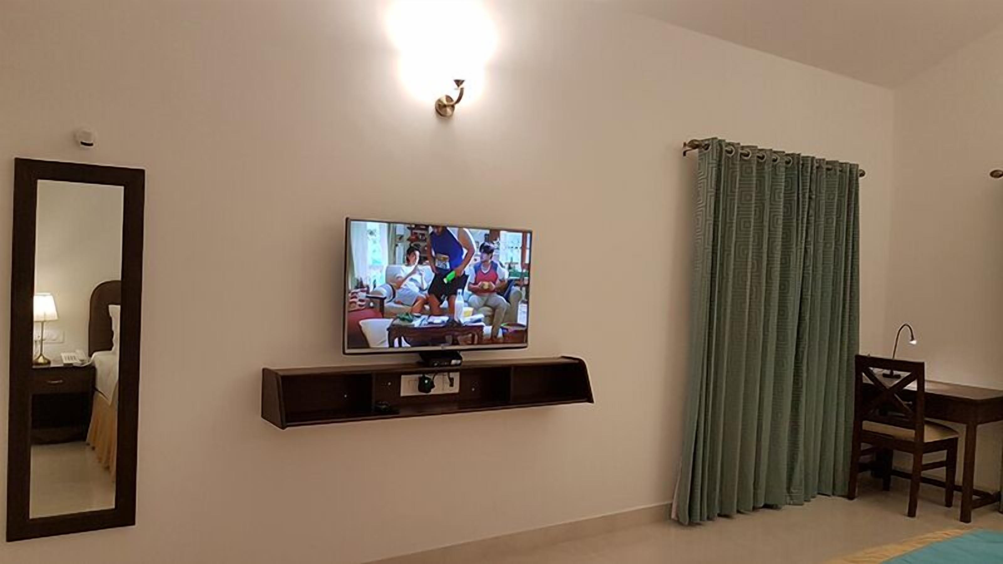 A living room with a flat-screen TV mounted on the wall, a wooden TV stand, a dining table with chairs, and a large mirror.