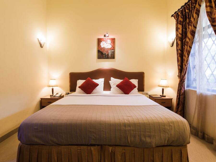 Habitación de hotel con una cama grande, dos mesitas de noche con lámparas, una obra de arte colgada en la pared y un ventanal con cortinas.