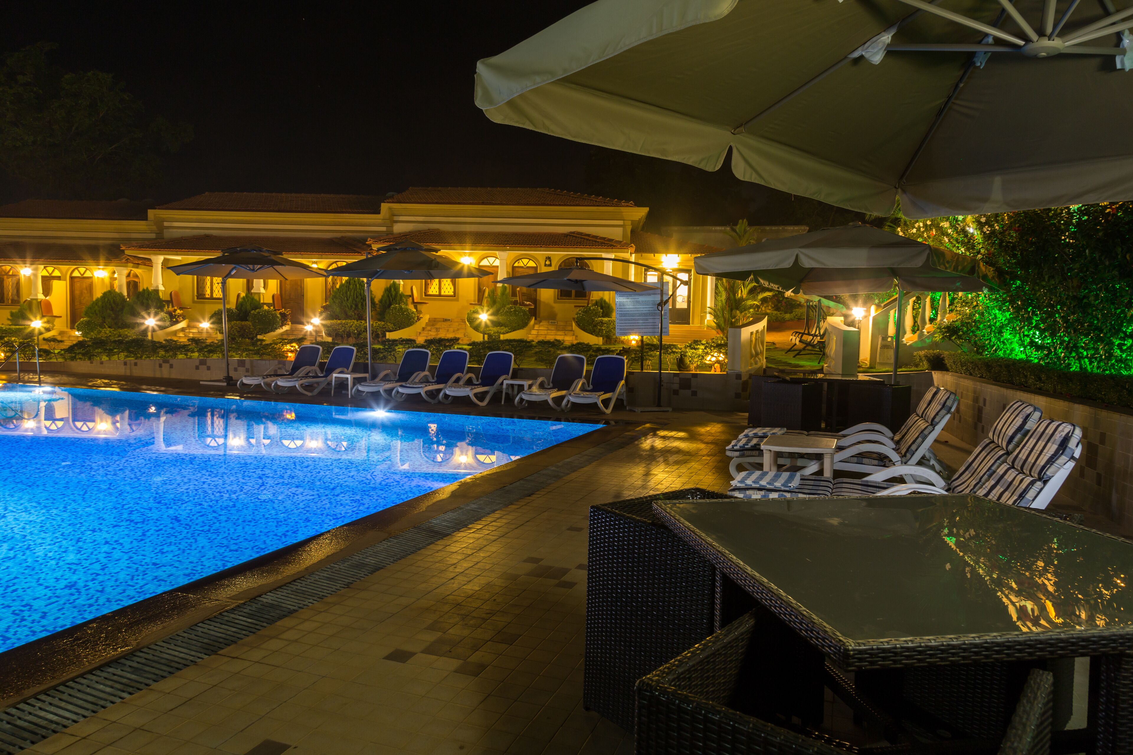 Ein Poolbereich bei Nacht mit Liegestühlen und Sonnenschirmen, einem Pool und einem Gebäude mit beleuchteten Fenstern.
