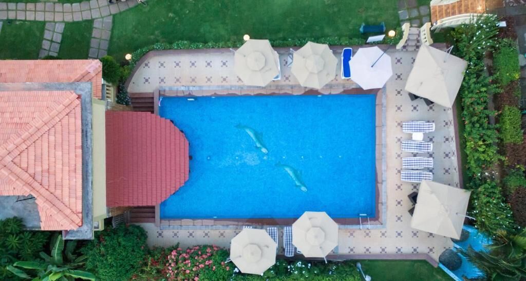 An aerial view of a swimming pool with a red-roofed building, surrounded by umbrellas and lounge chairs.