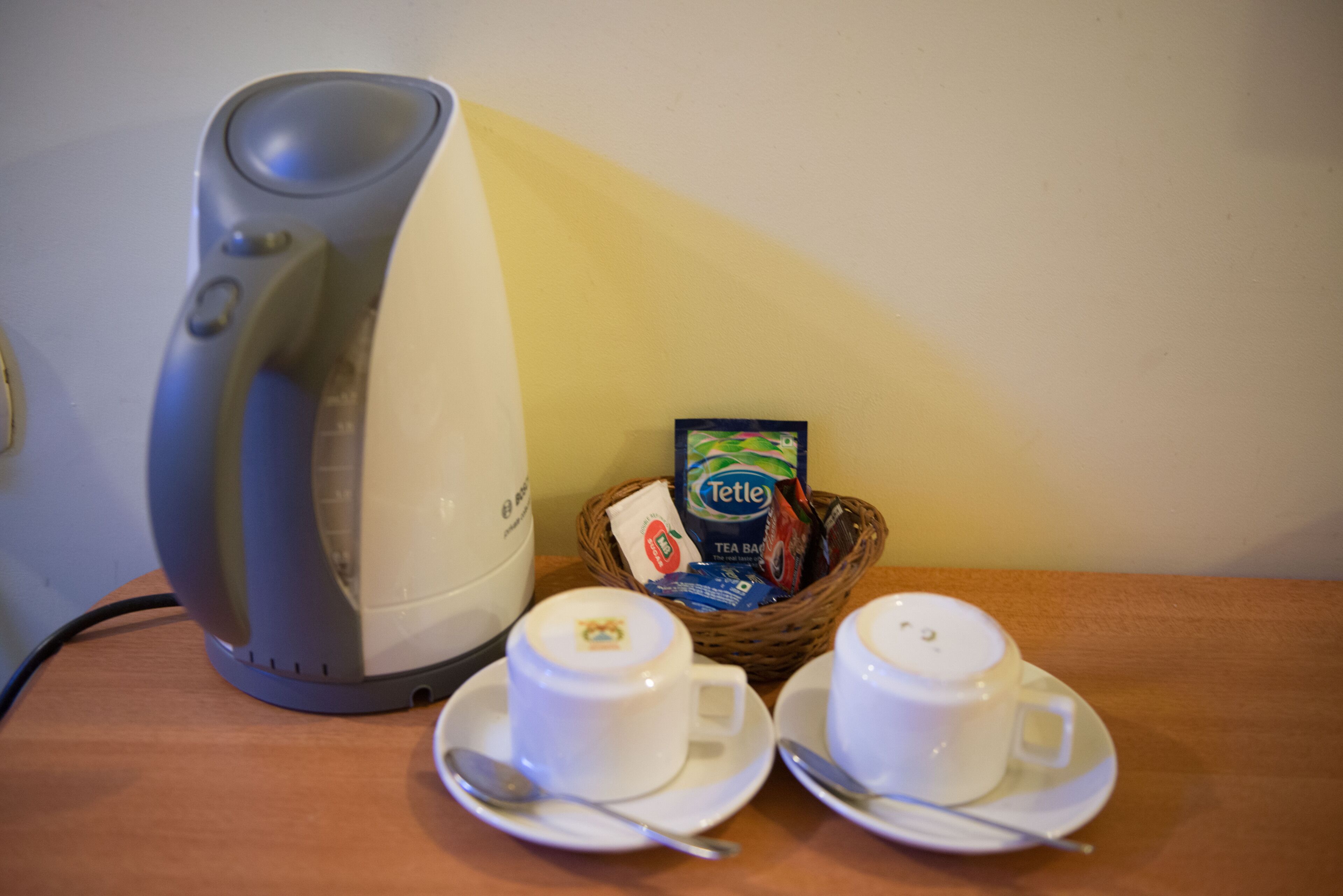 Une machine à café, deux tasses avec leurs soucoupes et un panier contenant des sachets de thé, le tout posé sur une table en bois.