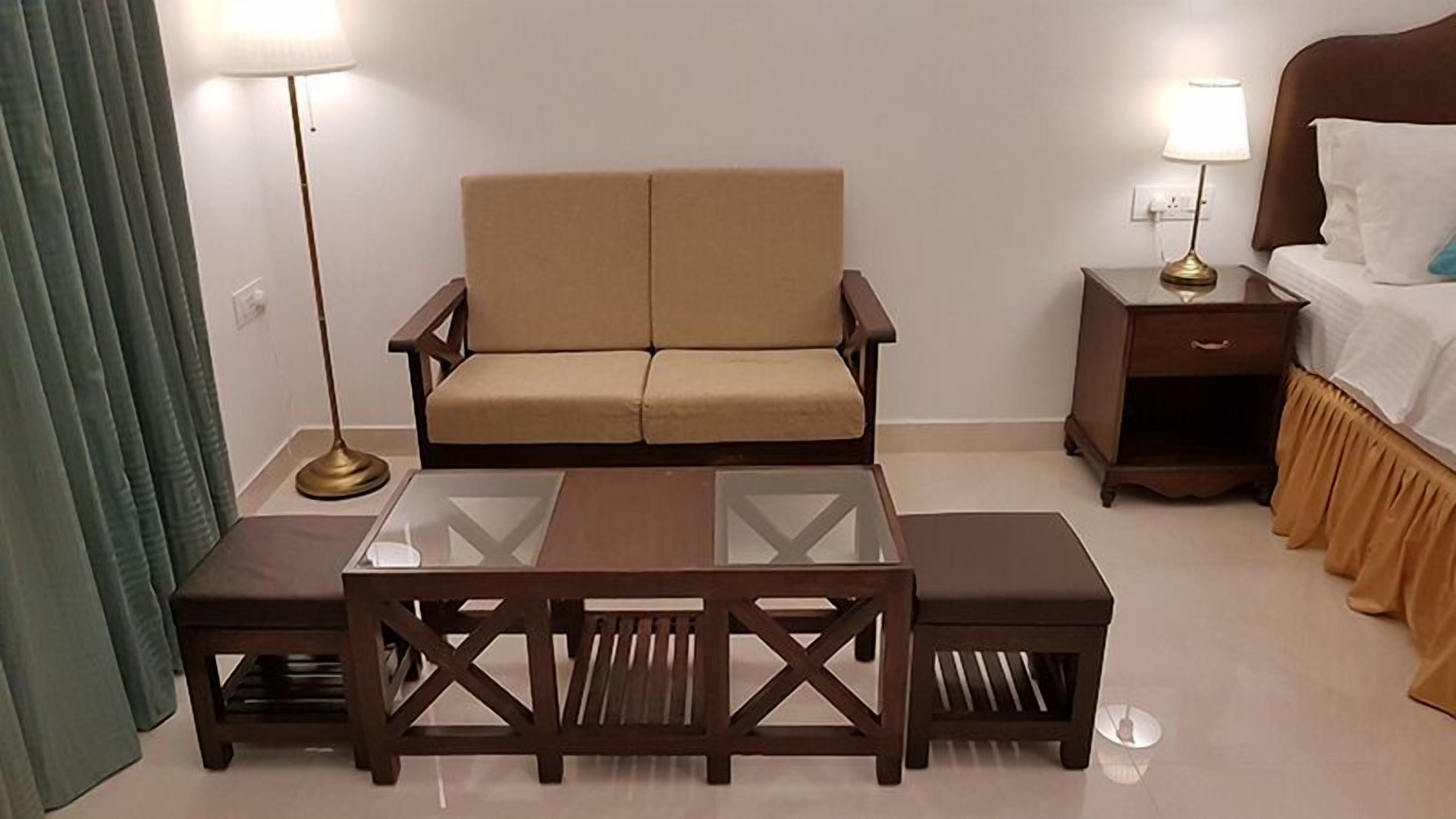 Ein Hotelzimmer mit einer Couch, einem Glastisch, einem Nachttisch mit Lampe und einem Bett mit weißem Kopfkissen.