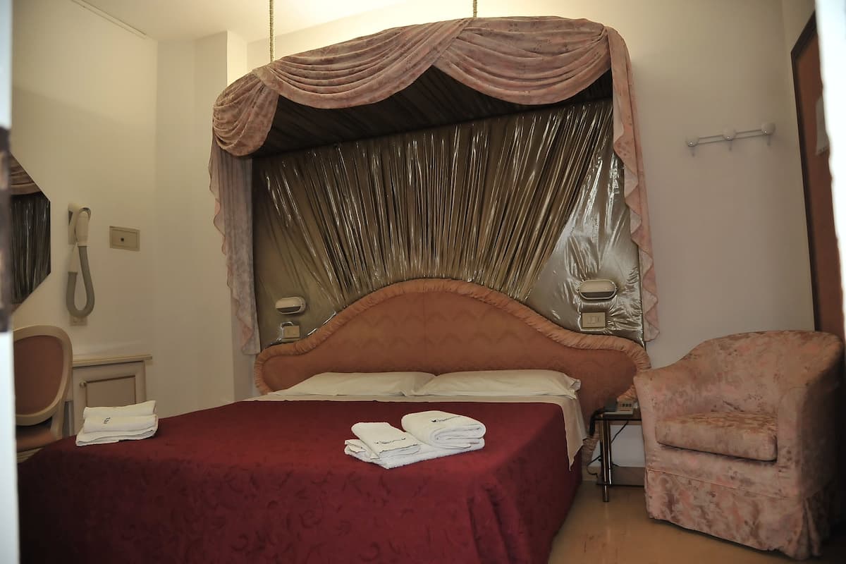 Een netjes opgemaakt bed met een rode sprei en witte kussens, een stoel en een nachtkastje met een lamp.