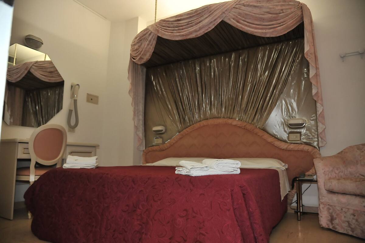Een opgemaakt bed met een rode sprei, een nachtkastje met een lamp en een stoel.