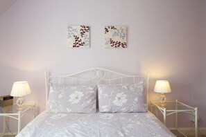 A bedroom with a white metal bed frame, floral bedding, two bedside tables with lamps, and two wall-mounted artworks. - Avonlea (Crieff)