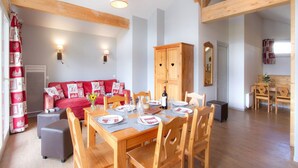 Uma sala de jantar com mesa de madeira posta para quatro pessoas, um sofá vermelho com almofadas estampadas em formato de coração e um armário de madeira.