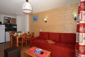 A cozy living room with a red sofa, wooden dining table, and chairs. There is a kitchen area with a microwave and a refrigerator. - Versatile Apartments in French Alps (Les Deux Alpes)