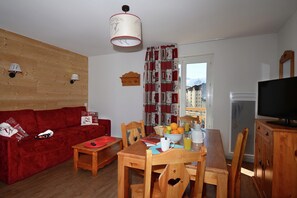 A cozy living room with a red sofa, wooden dining table, and a TV. - Versatile Apartments in French Alps (Les Deux Alpes)