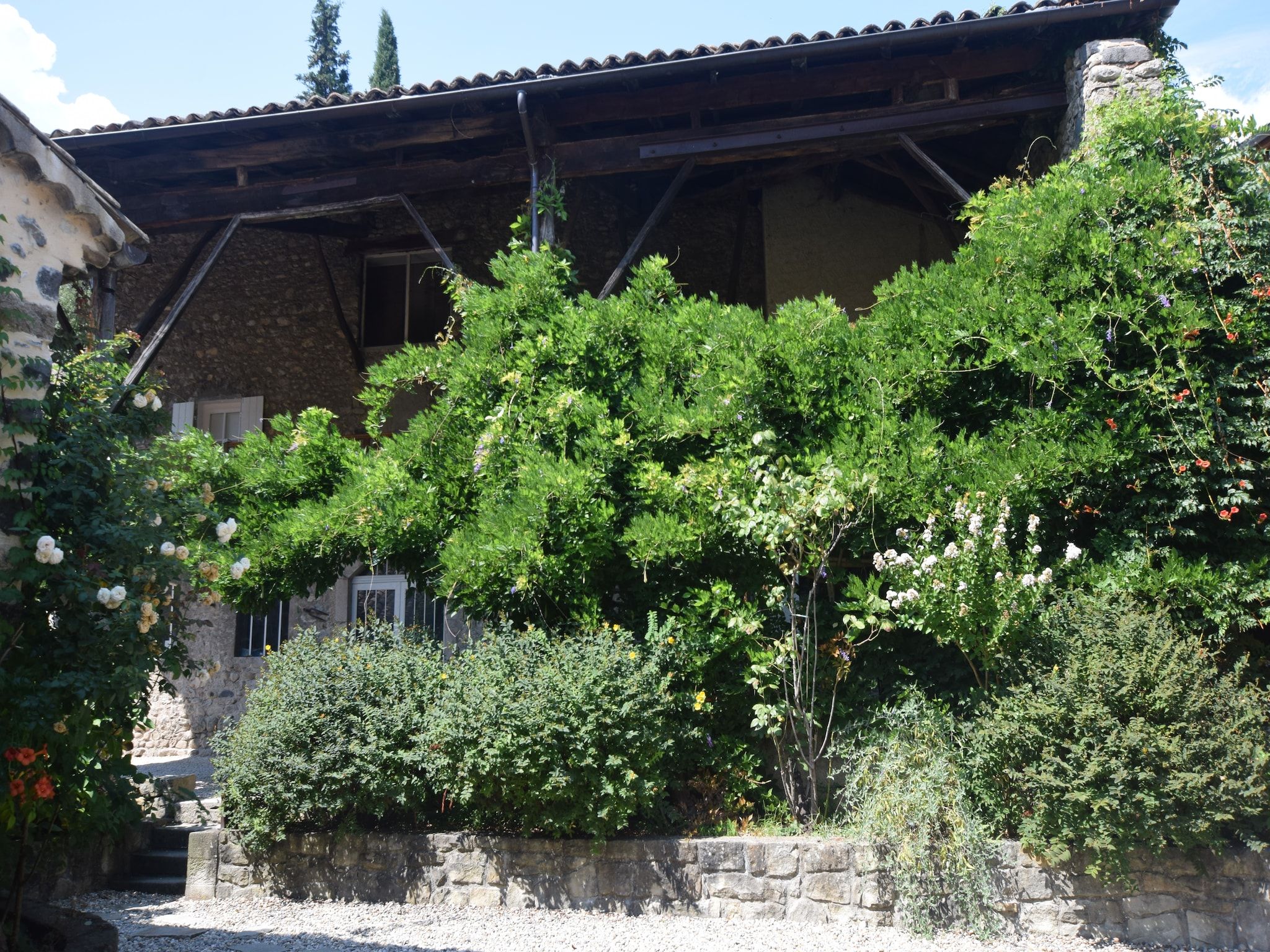 A stone building with a tiled roof, surrounded by lush greenery and flowering plants.