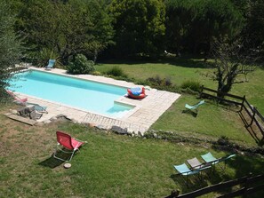 A rectangular swimming pool with a tiled deck, surrounded by a grassy area with trees. There are lounge chairs and a red chair near the pool. - Holiday Home in Ardèche with Pool & Garden (Pont-De-Labeaume)