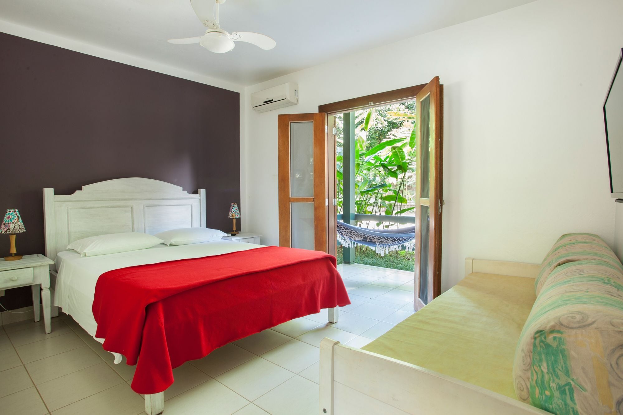 Une chambre avec un lit, une table de chevet, une couverture rouge, un hamac et une vue sur la verdure par une porte ouverte.