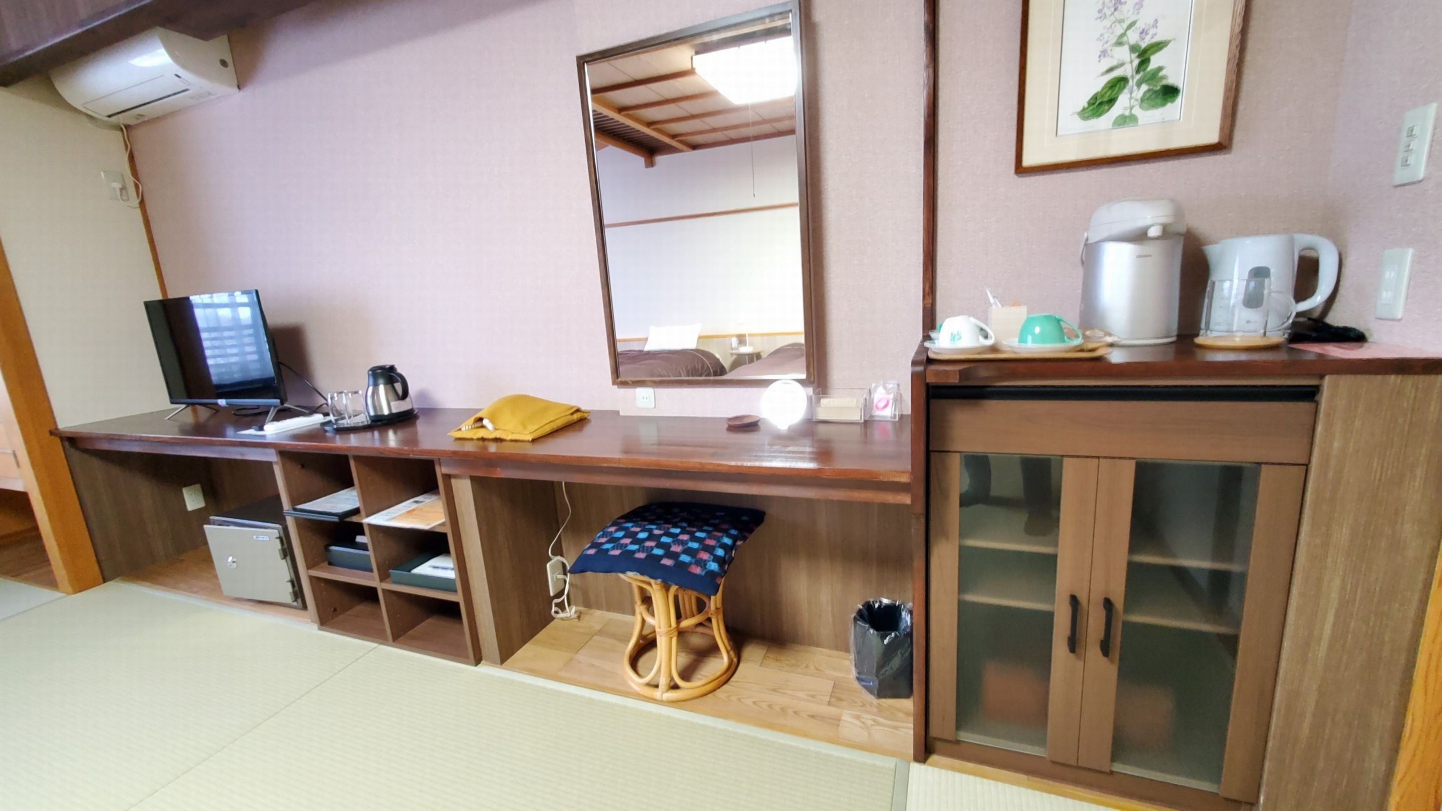 A hotel room with a wooden desk, a computer, a coffee maker, a tea set, and a cabinet with glass doors.