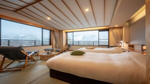 A spacious bedroom with a large bed, a seating area with chairs and a small table, and large windows offering a view of the city. - Kinosato (Yamagata)