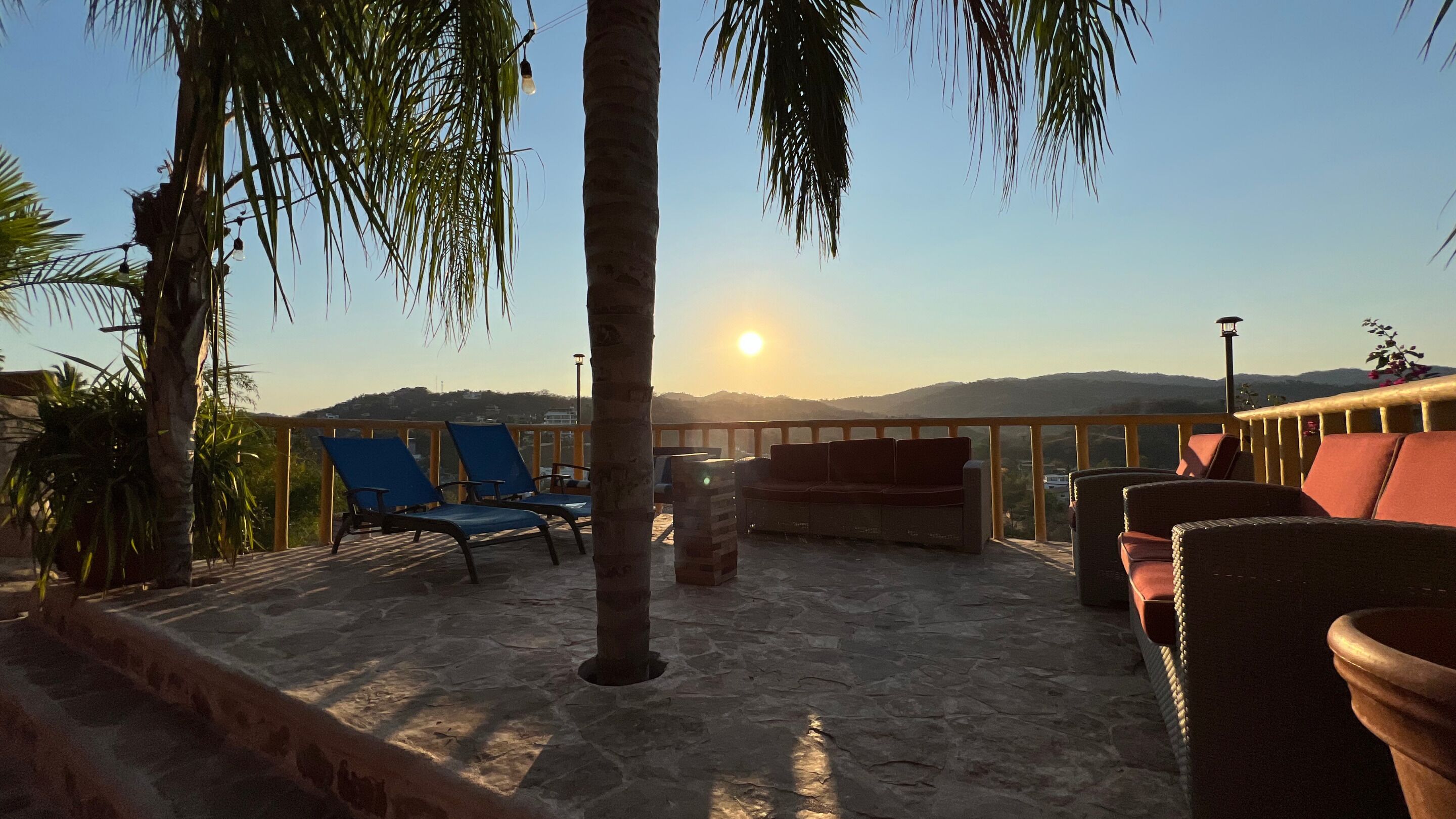 A terrace with stone flooring, blue and red lounge chairs, and a view of the sun setting over hills.