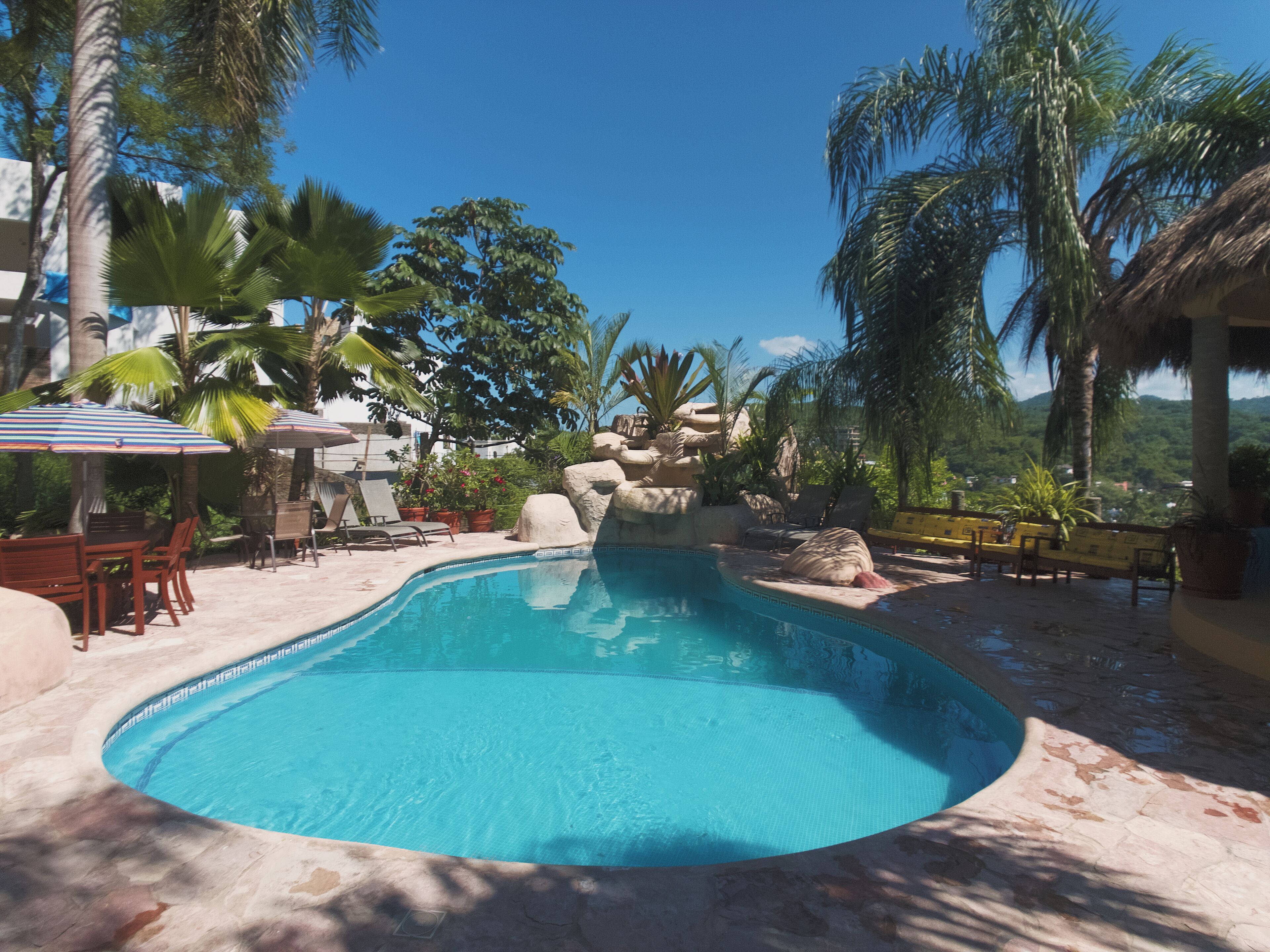 Ein Swimmingpool mit Liegestühlen und Sonnenschirmen, umgeben von Palmen und einem Bergpanorama.