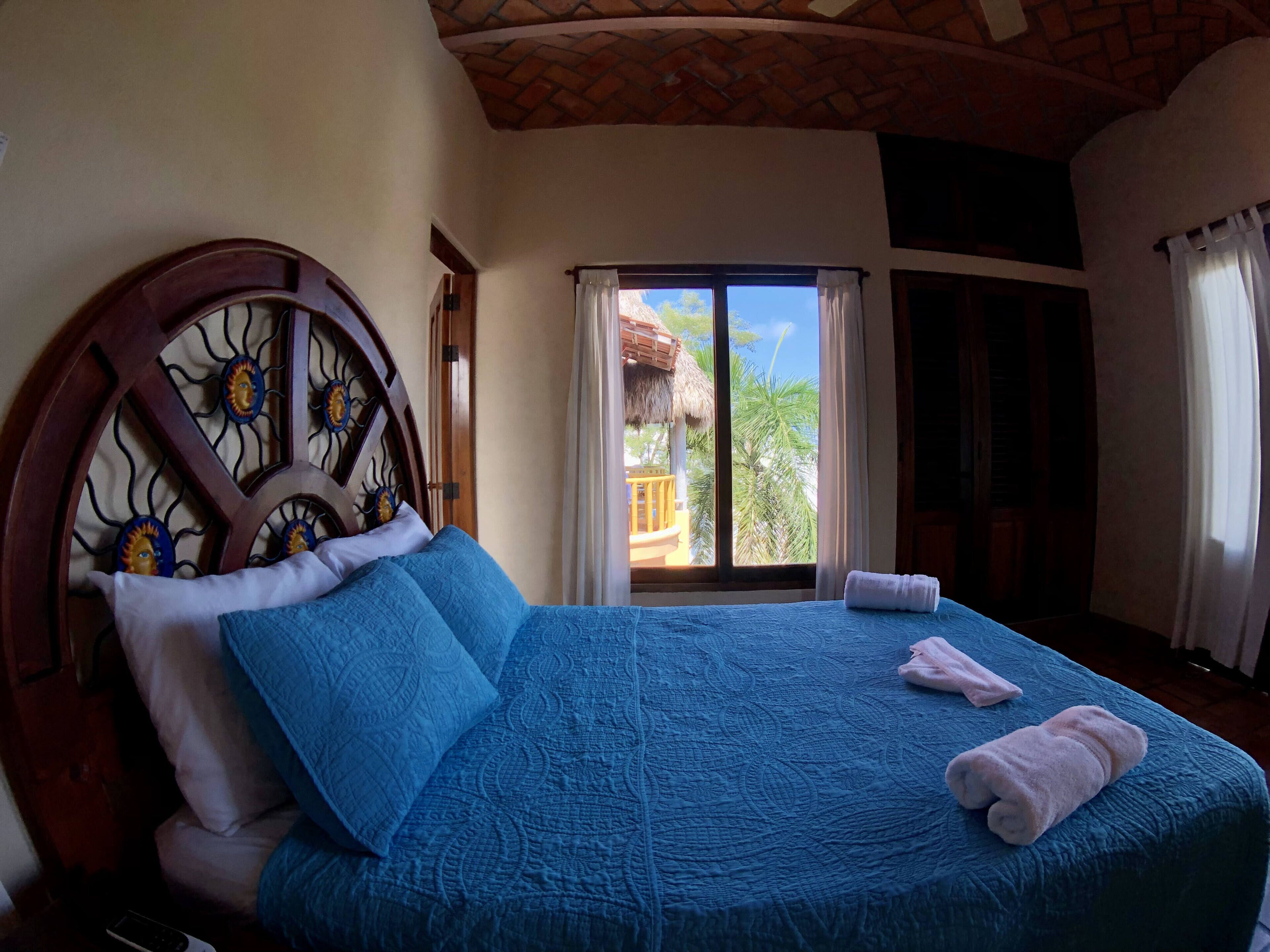 A bedroom with a large bed, a wooden headboard with sun motifs, and a view of a balcony with a thatched roof.