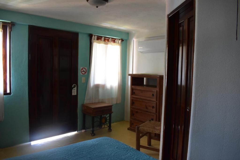 Une pièce aux murs de couleur turquoise, avec une commode en bois, une petite table, un lit et une porte sur laquelle est apposée une pancarte.