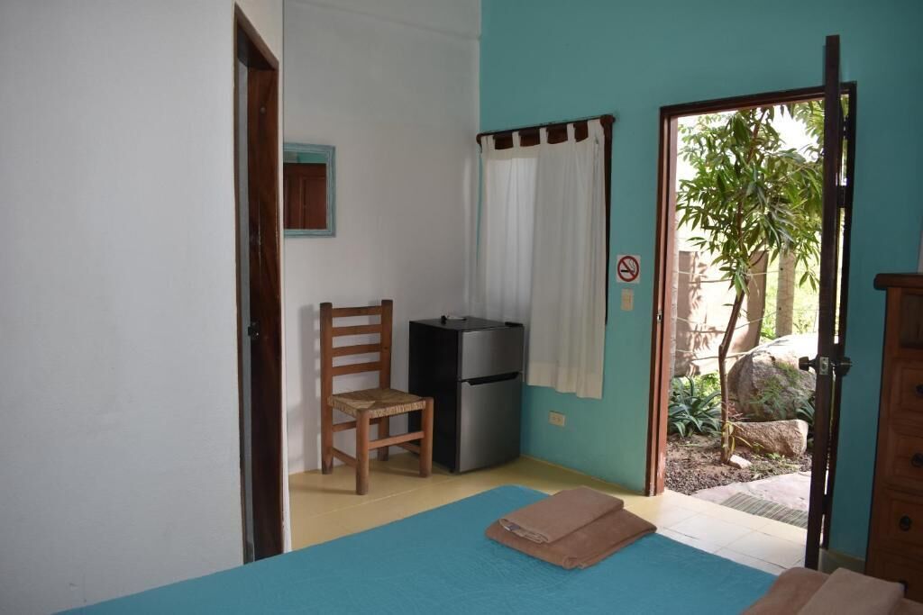A room with a bed, a chair, a small refrigerator, and a door leading to an outdoor area with plants.