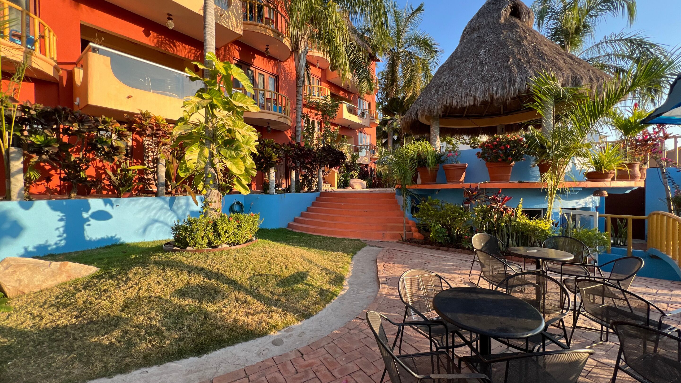 Un patio de hotel con mobiliario exterior, una estructura de techo de paja y exuberantes plantas tropicales.