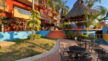 Un patio de hotel con mobiliario exterior, una estructura de techo de paja y exuberantes plantas tropicales.