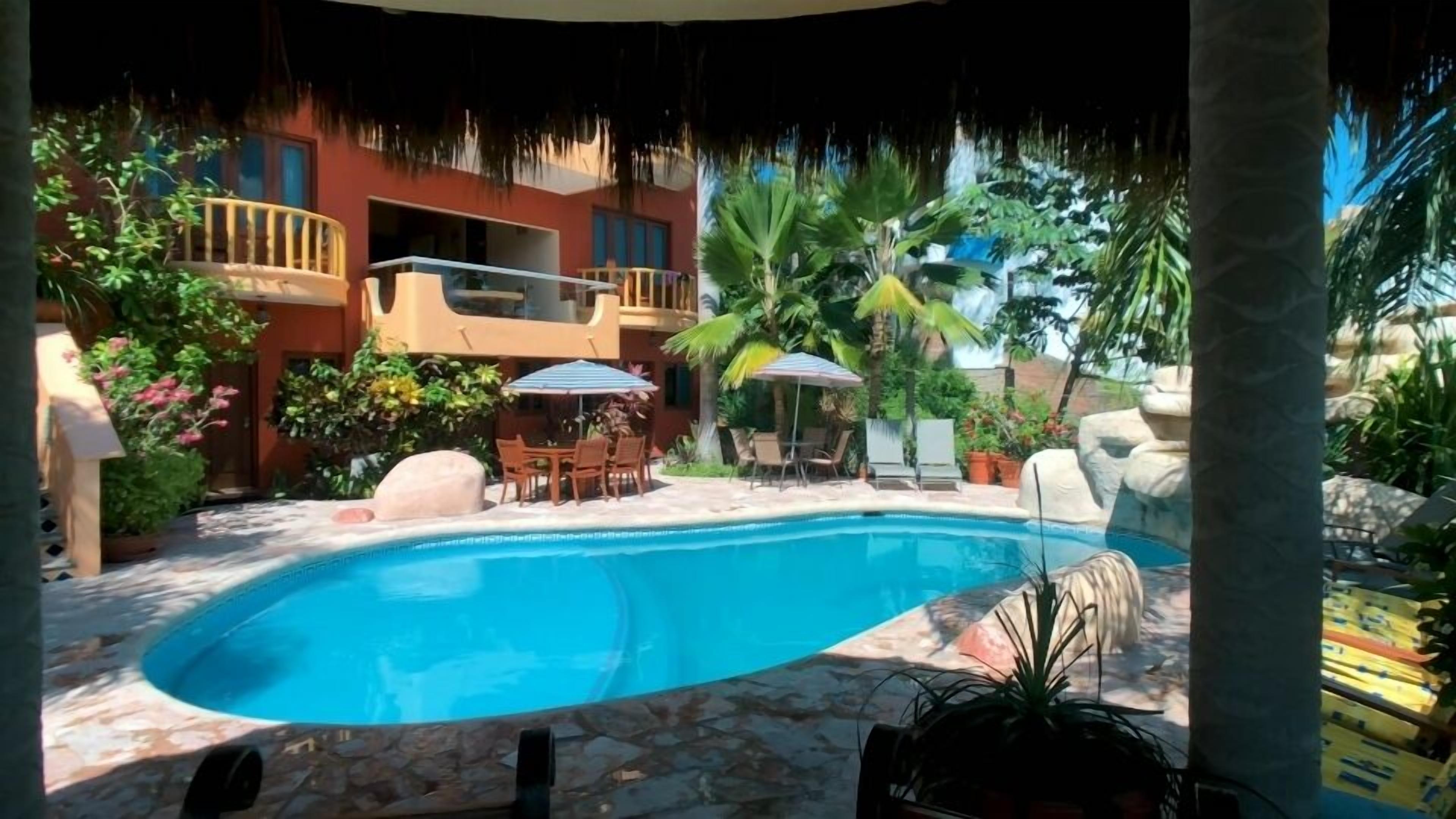 A swimming pool with lounge chairs and umbrellas, surrounded by tropical plants and a building with balconies.