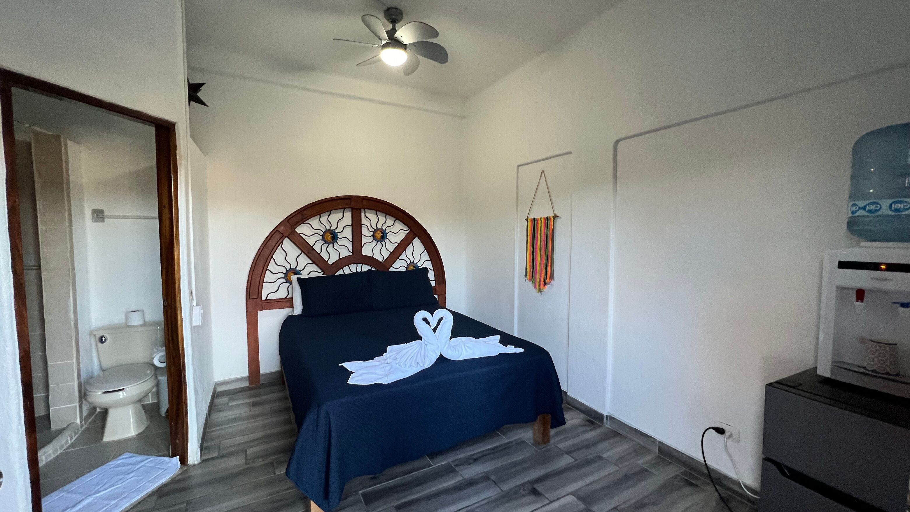 A bedroom with a bed, a ceiling fan, a water dispenser, and a bathroom with a toilet and sink.