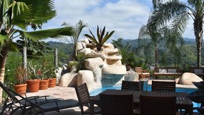 Un espace extérieur avec piscine, des chaises longues et des tables, entouré de plantes en pot et d’une formation rocheuse.