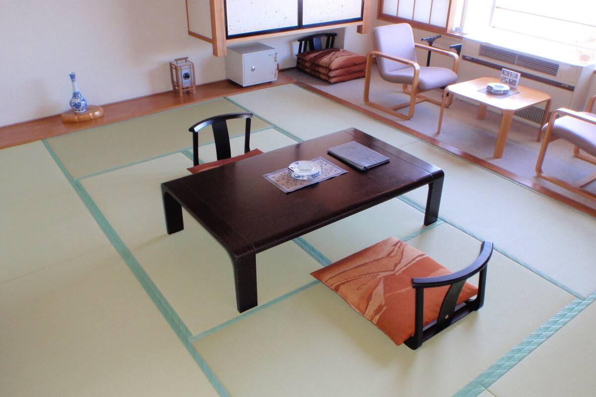 A tatami mat room with a low table, two chairs, a small heater, and a TV on a stand.