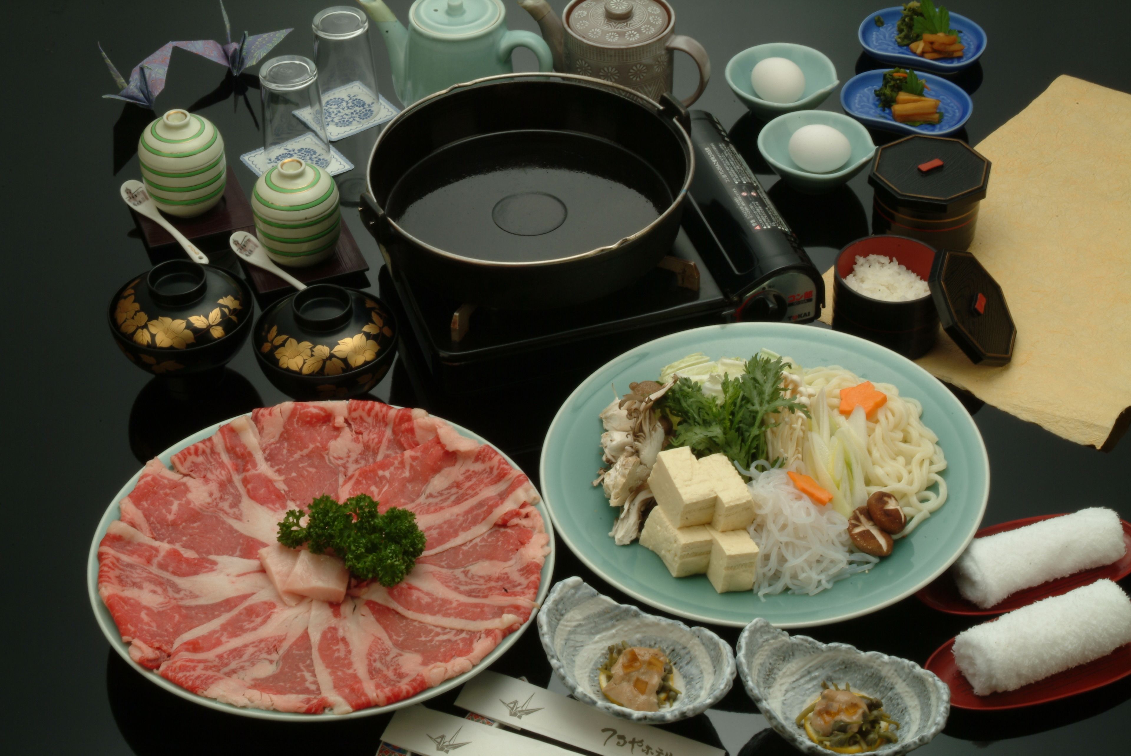 A table with various dishes, including a plate of raw meat, a bowl of noodles, and several small dishes with different ingredients.