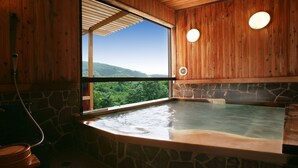 Una cabaña de madera con un manantial de aguas termales en una bañera de piedra, un banco de madera y vistas a una montaña boscosa.