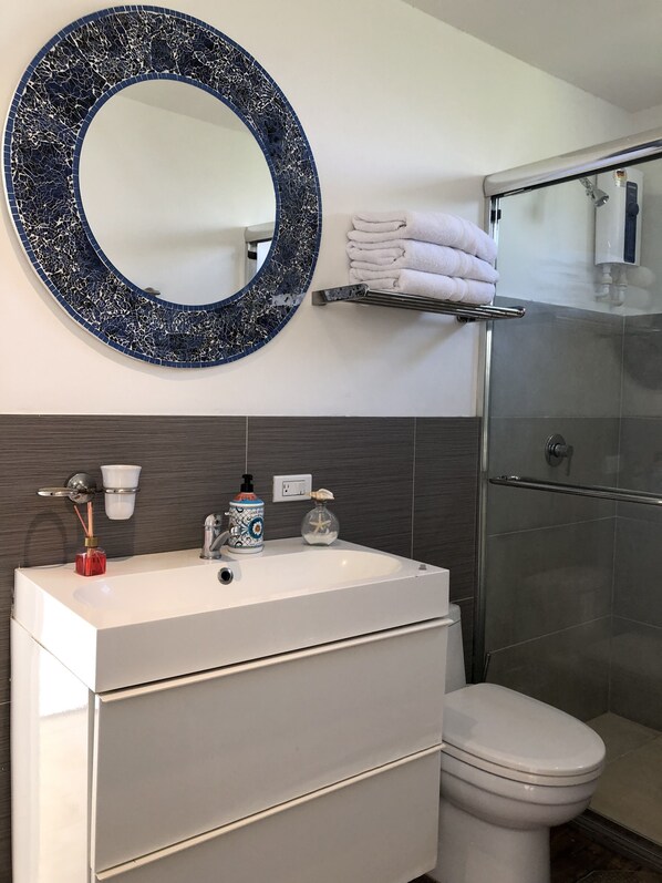 A bathroom with a white sink, a round mirror with a blue mosaic frame, a towel rack with folded towels, a glass shower enclosure, and a toilet.