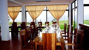 A restaurant with a long wooden table set for dining, large windows with curtains, and a view of a green landscape. - Hosteria Farallon (Sevilla Don Bosco)