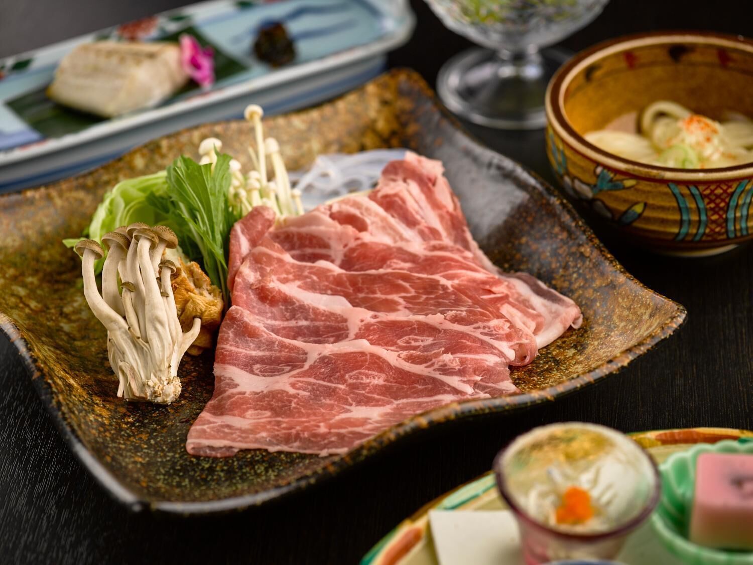 A plate with slices of meat, mushrooms, and greens, accompanied by a bowl of soup and a small dish with a pink item.