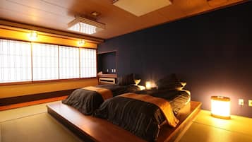 A traditional Japanese-style room with tatami flooring, two beds with dark bedding, a low wooden platform, and a large window with vertical blinds.