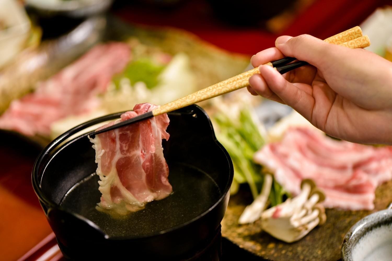 A person holding a piece of meat over a black pot, likely a hot pot or broth, with chopsticks.