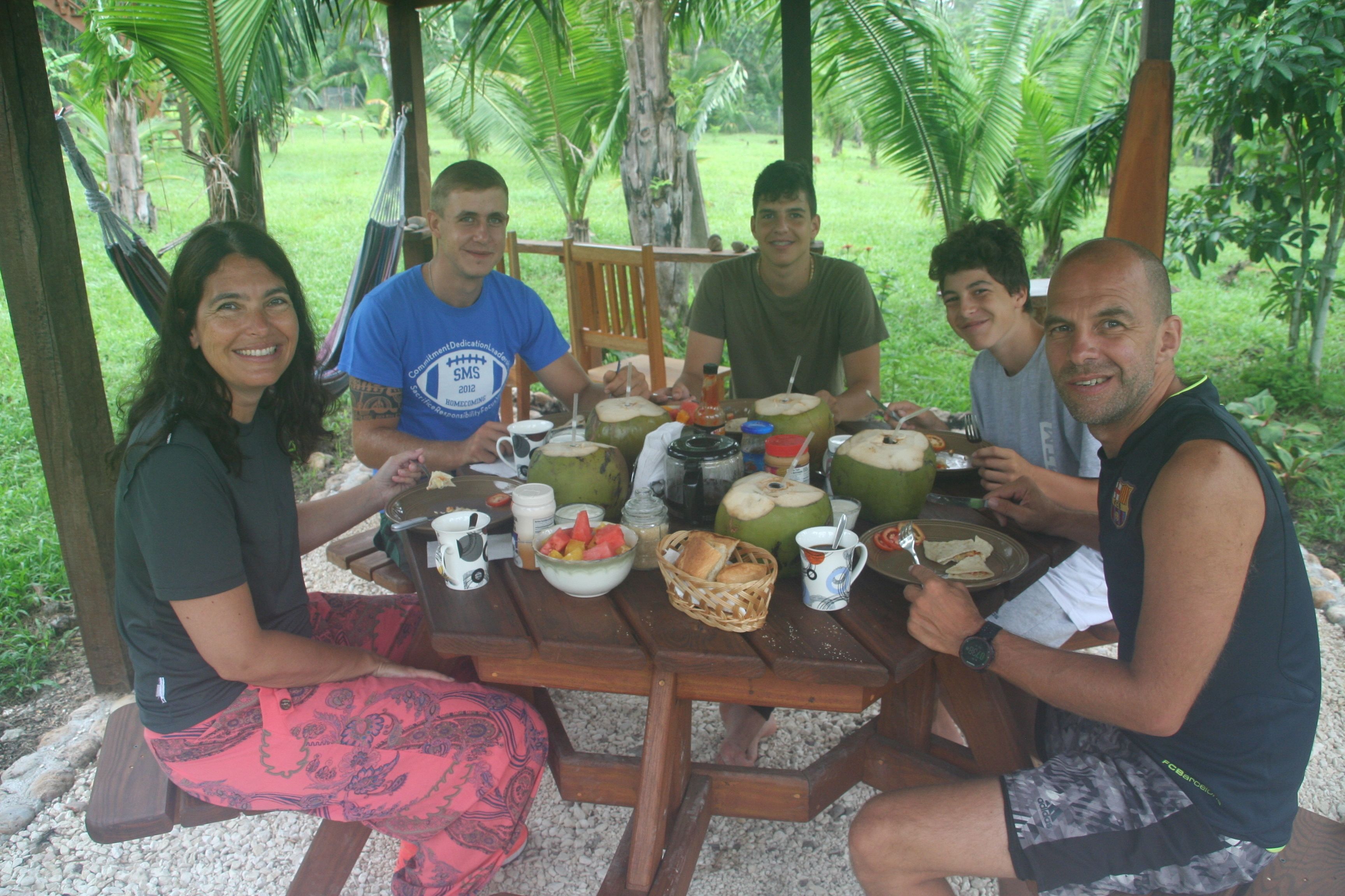 A group of people sitting at a wooden table outdoors, with a picnic setup including coconuts, bread, and fruit.