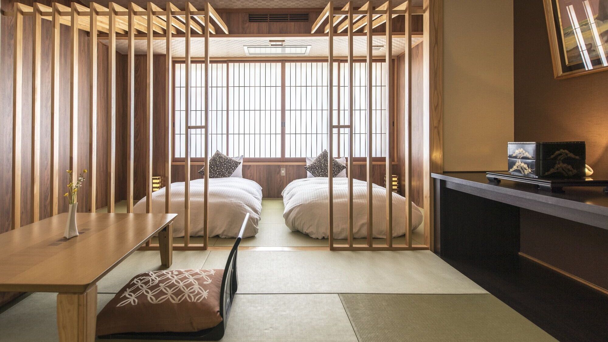 A traditional Japanese-style room with tatami flooring, two beds with pillows, a low table, and a desk with a computer.