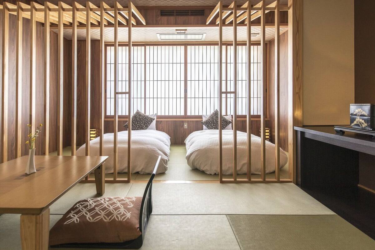 Um quarto tradicional japonês com tatames, duas camas com travesseiros, uma mesa baixa e uma escrivaninha com um computador.