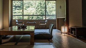 Perinteinen japanilainen huone, jossa on tatamimatto, matala pöytä tyynyineen ja suuri ikkuna, josta avautuu näkymä vehreään maisemaan.