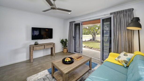 A living room with a ceiling fan, a flat-screen TV, a wooden console table, a coffee table with fruits and a chess set, a blue sofa with a yellow throw, and a view through a glass door. - 12th Tee Bed and Breakfast (Bargara)