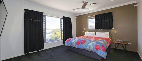 A bedroom with a bed, a ceiling fan, a window with curtains, and a small table with a lamp.