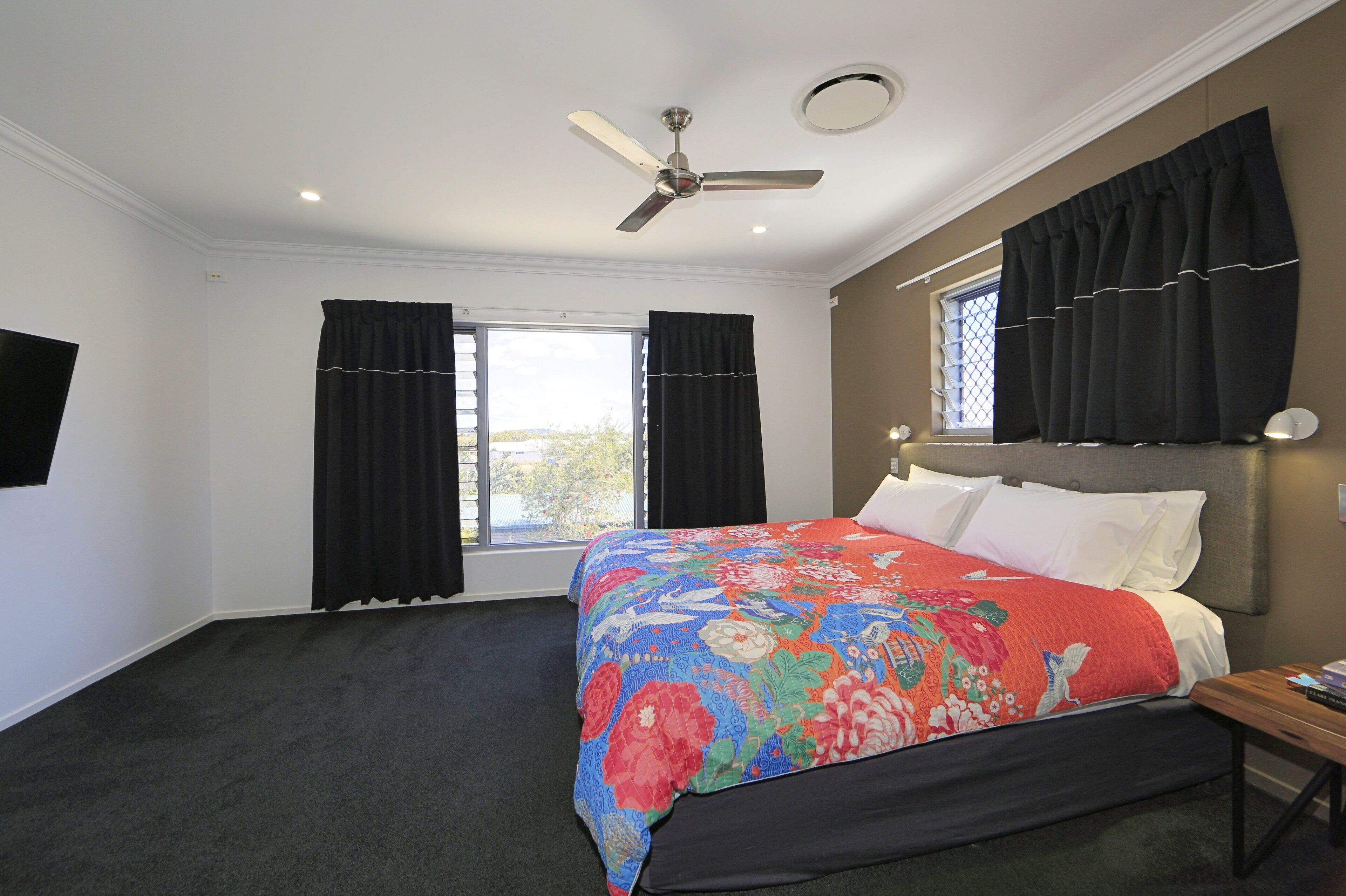 A bedroom with a bed, a ceiling fan, two windows with black curtains, and a nightstand with a lamp.