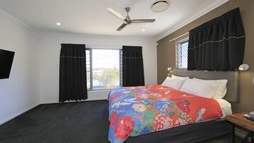 A bedroom with a bed, a ceiling fan, two windows with black curtains, and a nightstand with a lamp.