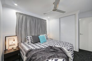Un dormitorio con cama, mesita de noche, un cuadro enmarcado, ventilador de techo y una puerta.