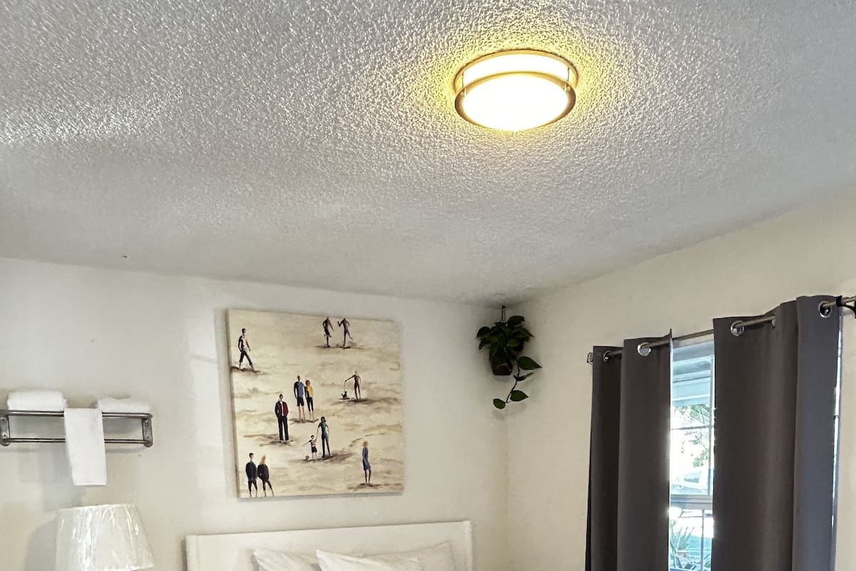 Un dormitorio con cama, mesita de noche, lámpara, un cuadro, ventana con cortinas y luz de techo.