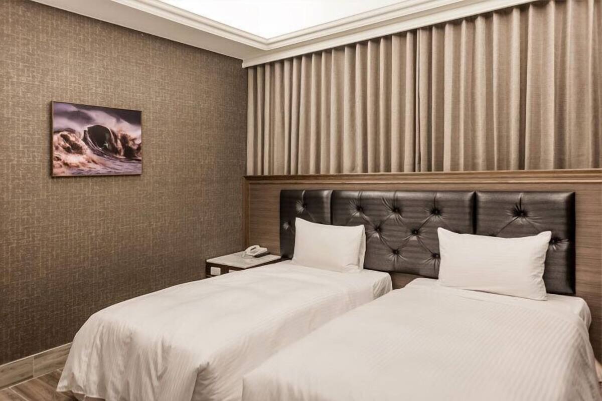 A hotel room with two beds, a headboard, a nightstand, and a painting on the wall.
